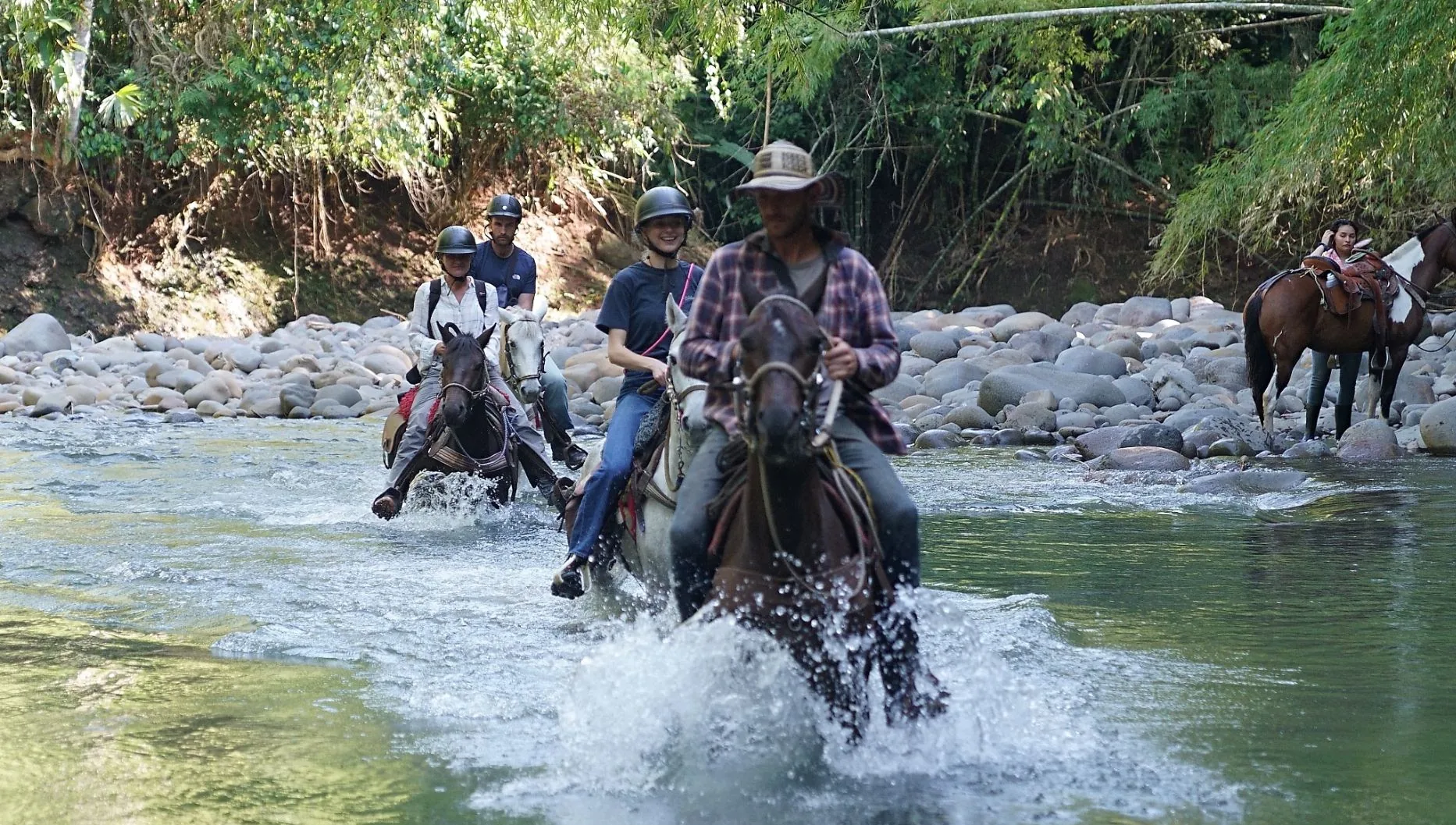 Riders and horses wading a wide river between boulders and jungle.